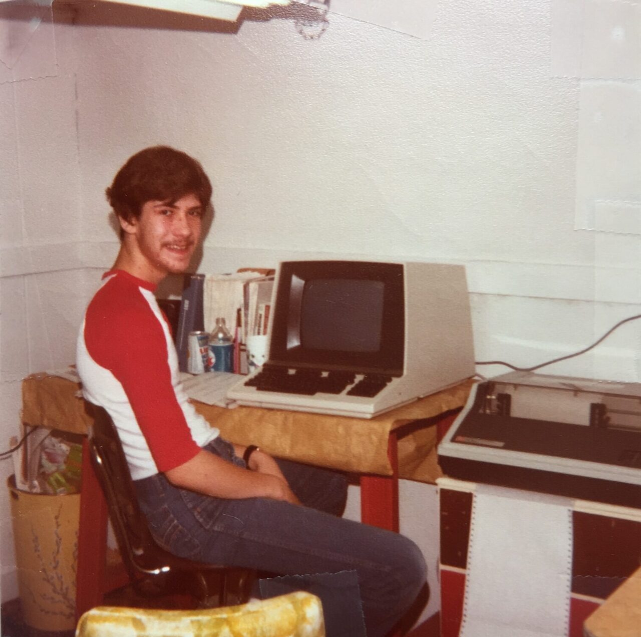 A teenage Doug in a red and white baseball shirt, sitting at a desk beside a computer terminal and a printer, smiling at the camera.